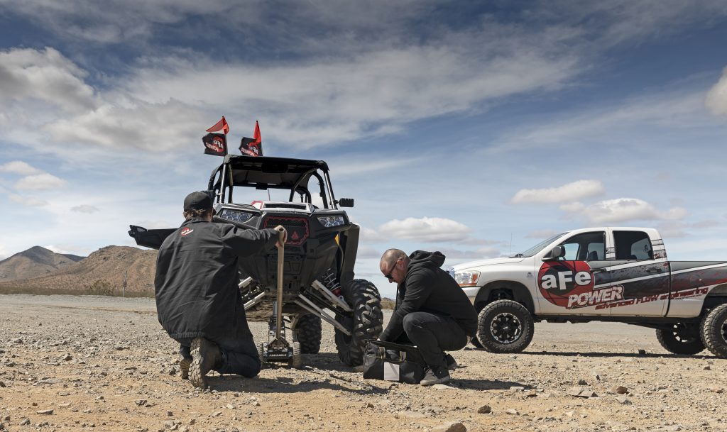 Two men kneeling on desert ground fixing up side-by-side Polaris in Ocatillo Wells offroading Southern California desert excursion with big white aFe POWER branded truck in back