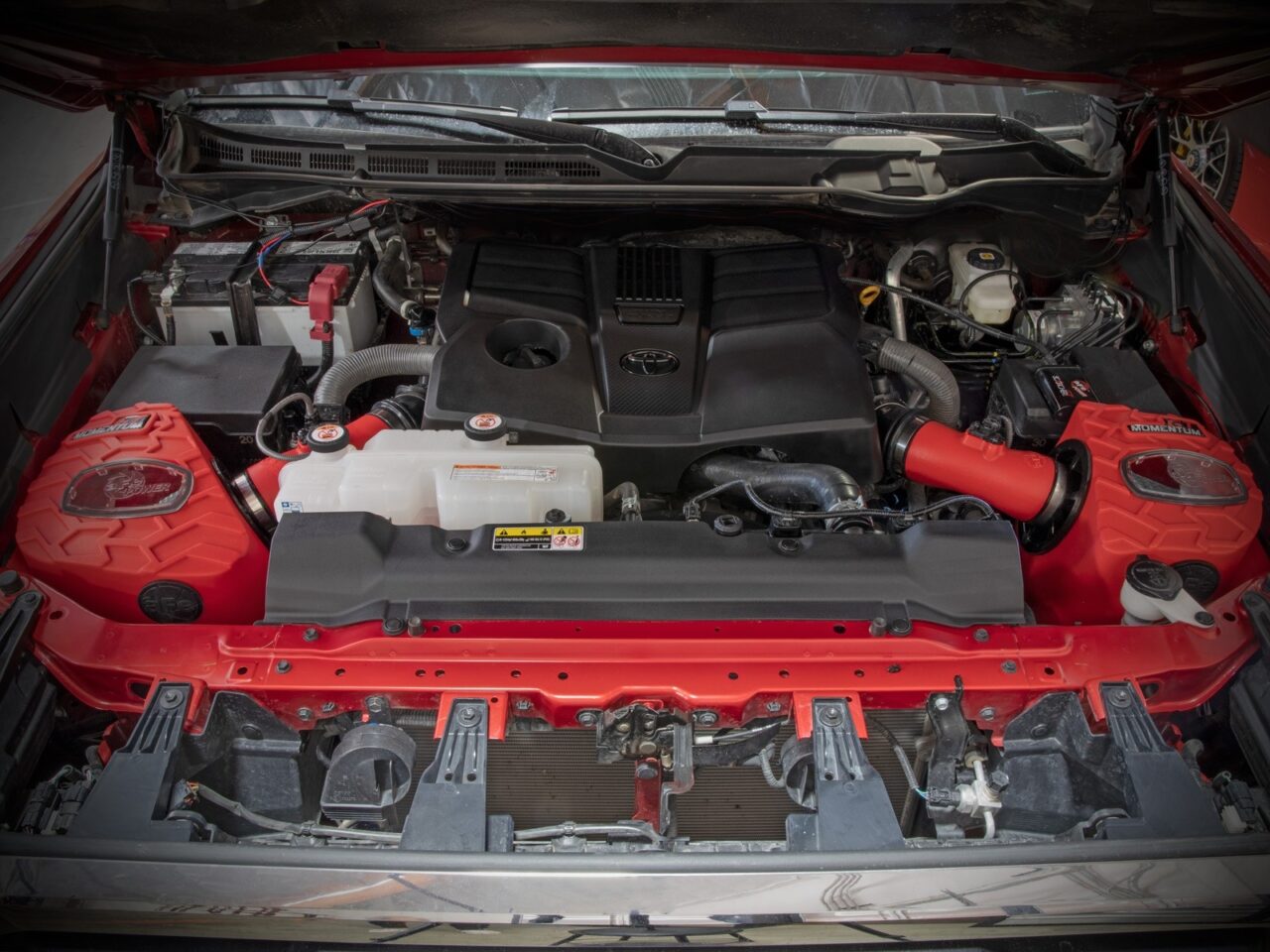 Two bright red airbox cold air intakes with tire tread design and with clear sight windows and red tubes connected to V6 3.4L twin-turbo engine on Red 2022 3rd Gen Toyota Tundra